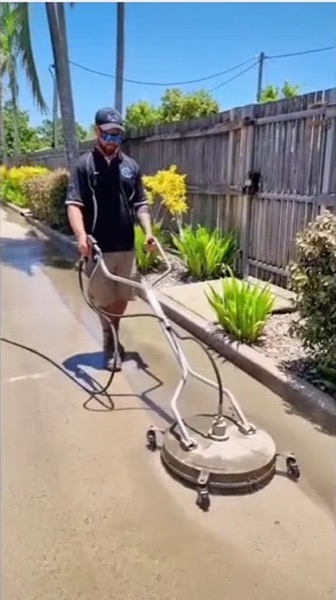 A worker walking a surface cleaner down a driveway beside a tropical garden bed