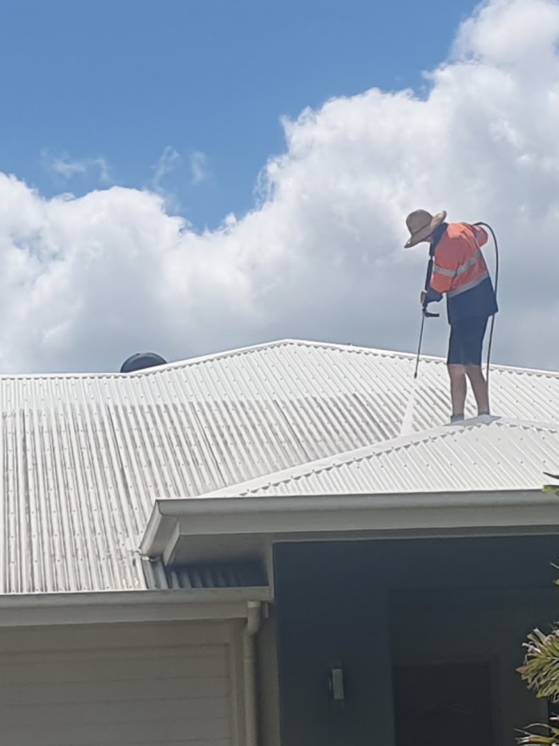 A worker in hi-vis standing on a white metal roof, washing it under a cloudy Mackay sky