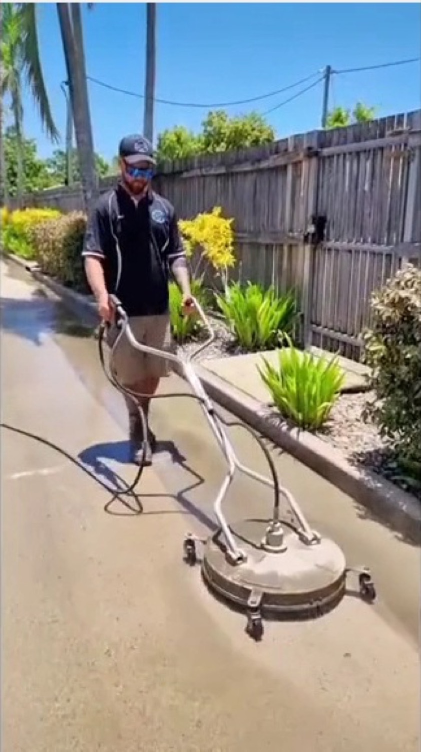 A worker walking a surface cleaner along a driveway beside a tropical garden bed